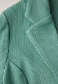 Muntgroene ribgebreide blazer met een reverskraag en prominente zoomdetails, die de textuur van de stof en het gestructureerde ontwerp benadrukken.