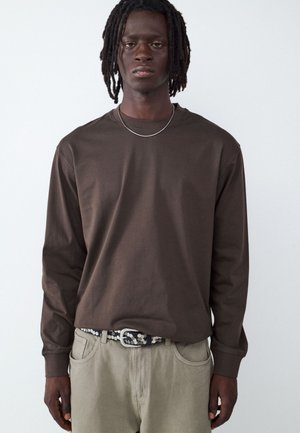 Young man with medium-dark skin and dreadlocks wearing a brown long-sleeve shirt, light beige pants, a studded belt, and a thin necklace.