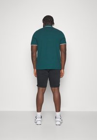 Turkusowa koszulka polo z białymi akcentami, zestawiona z czarnymi szortami. Model zwrócony plecami, w białych trampkach. Gładki materiał, luźny krój.