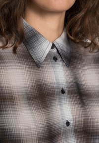 Karierte Bluse in Grautönen und Weiß, mit einem knöpfbaren Kragen und schwarzen Knöpfen, die eine weiche, leichte Stoffstruktur zeigt.