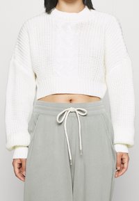 Weißer, kurzer Strickpulli mit einem Zopfmuster, ausgestattet mit langen Ärmeln und gerippten Bündchen, kombiniert mit hellgrauen Jogginghosen mit Kordelzug.