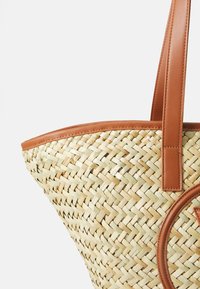 Vävd stråväska med en ljusbrun läderkant och handtag. Har en rund läderdetalj på sidan. Texturerad yta med naturliga mönster.