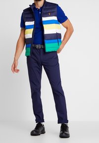 Uppblåsbar väst med marinblå, vit, grön, blå och gul horisontella ränder; bärs över en blå polo; i kombination med marinblå byxor och svarta skor.