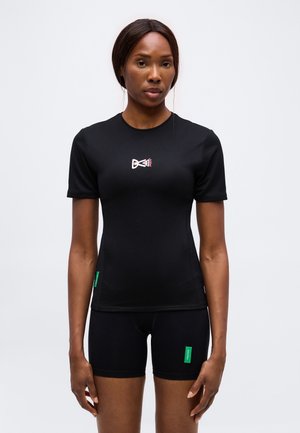 Femme portant un t-shirt noir ajusté à manches courtes avec un petit motif sur la poitrine et un short noir assorti avec des étiquettes vertes.