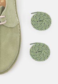 Světlé zelené semišové boty s béžovými tkaničkami, se dvěma stočenými zelenými tkaničkami zdobenými černými tečkami po stranách.