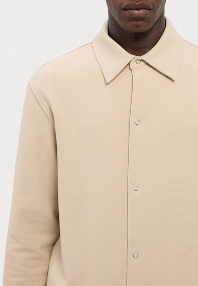 Beige kraevi särk pehmest kangast, millel on nööpide ja hõbedase riistvaraga esiosa ning sile tekstuur.