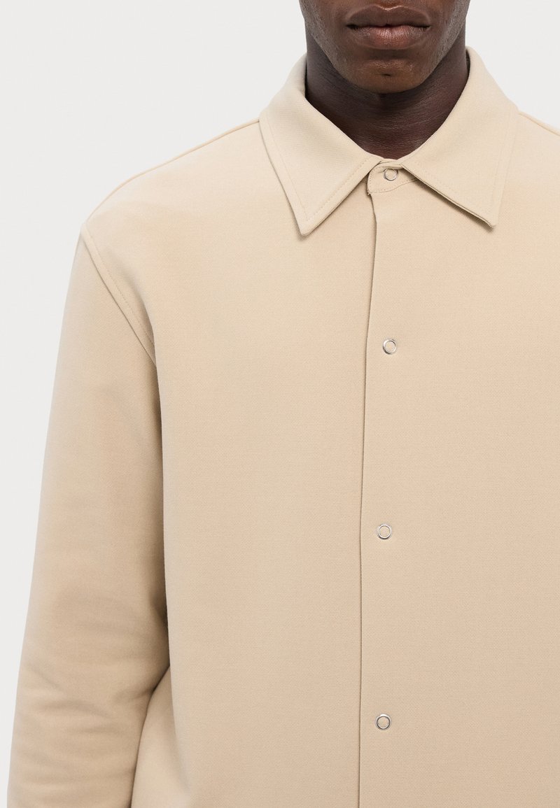 Beige overhemd met kraag, gemaakt van zacht materiaal, met een knoopsluiting aan de voorkant en zilveren hardware met een gladde textuur.
