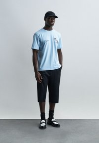 Άνδρας που φοράει ανοιχτό μπλε μπλουζάκι με σχέδιο, μαύρο καπρί παντελόνι, μαύρο καπέλο, μαύρες κάλτσες και μαύρα-άσπρα μοκασίνια, στέκεται μπροστά σε ένα απλό φόντο.