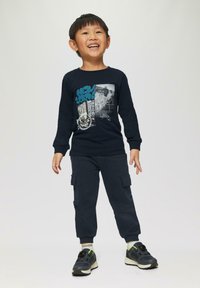 Marineblauw long sleeve shirt met grafische print, gecombineerd met marineblauwe cargobroek met zijzakken en zwarte schoenen met groene accenten.