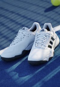 Pár bielych športových topánok Adidas s čiernou podrážkou a pruhmi položených na modrom tenisovom kurte vedľa žltej loptičky.