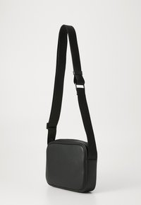 Sac à bandoulière rectangulaire noir avec une sangle en tissu large et réglable, et une fermeture éclair, présenté sur un fond clair neutre.