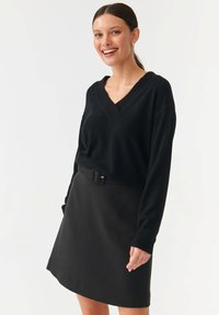 Schwarzer V-Ausschnitt-Pullover mit langen Ärmeln, kombiniert mit einem schwarzen Rock mit Gürtel. Beide Teile haben eine glatte Textur und ergeben ein stimmiges Outfit.