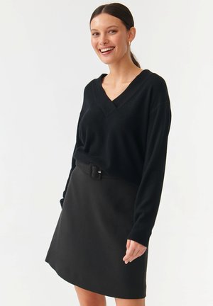 Zwarte v-hals trui met lange mouwen, gecombineerd met een zwarte rok met een riem. Beide hebben een soepele textuur, waardoor een samenhangende outfit ontstaat.