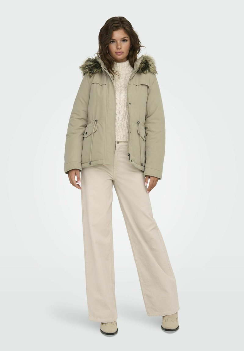Khaki Parka mit Kapuze aus Kunstfell, zwei vorderen Taschen und Kordelzug in der Taille. Getragen über einem cremefarbenen Pullover mit Zopfmuster und beigen, weiten Hosen.