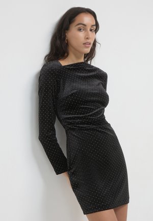 Schwarzes Samtkleid mit langen Ärmeln, verziert mit kleinen goldenen Punkten, tailliert geschnitten und mit einem runden Ausschnitt, der direkt oberhalb des Oberschenkels endet.