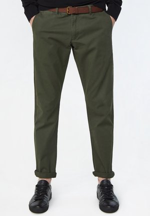 Man draagt opgerolde olijfgroene broek, zwarte riem, zwarte sneakers, en heeft handen in de zakken tegen een effen witte achtergrond.