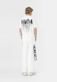 Camiseta blanca de manga corta con un gráfico de goteo de pintura negra en la espalda. Combinada con pantalones blancos y zapatos gruesos de colores claros.