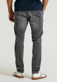 Hombre con jeans grises desgastados y zapatillas blancas de pie contra un fondo claro y liso, con las manos en las caderas.