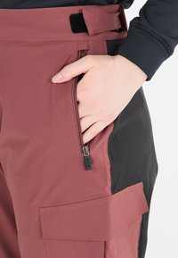 Mörkröda och svarta cargobyxor med en sidogenomskinlig ficka; tillverkade av ett slitstarkt, slätt material, med ett kardborreband och detaljfickor.