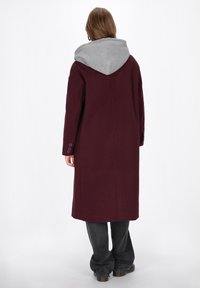 Cappotto bordeaux con cappuccio grigio, maniche lunghe e polsini abbottonati. Il design presenta una vestibilità rilassata e una silhouette fino al ginocchio.