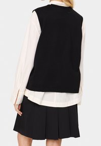 Czarny dzianinowy bezrękawnik nałożony na biały koszulowy shirt, w zestawieniu z plisowaną czarną spódnicą. Prosty fason z prążkowanymi krawędziami.