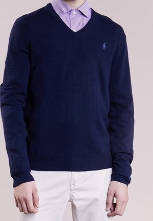 Man draagt een marineblauwe V-hals trui over een paars geruit overhemd, gecombineerd met een witte broek, staat met ontspannen armen.