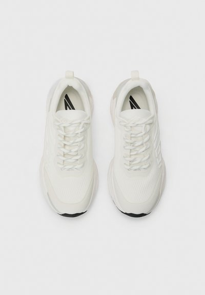 Weiße Sportschuhe mit einer strukturierten Oberseite, Schnürsenkeln und einer gepolsterten Sohle, mit einem schwarzen Gummiakzent an der Unterseite.