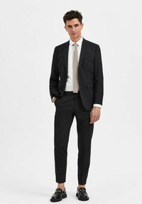 Svart dress med slim-fit design, med en enkel knapp på jakken. Kombinert med et lyse farget slips og glansede svarte sko.