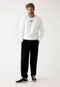 Sweat-shirt blanc avec logo noir, accompagné de joggers noirs et de baskets noires. Design simple et coupe décontractée, adapté pour une tenue décontractée.