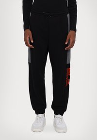 Pantaloni tuta neri con pannelli laterali grigi, vita elastica, cordoncino regolabile e dettaglio del logo rosso sulla coscia sinistra. Abbinati a sneaker bianche.