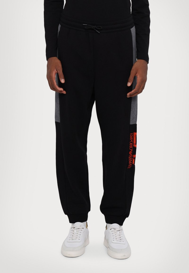 Pantaloni tuta neri con pannelli laterali grigi, vita elastica, cordoncino regolabile e dettaglio del logo rosso sulla coscia sinistra. Abbinati a sneaker bianche.