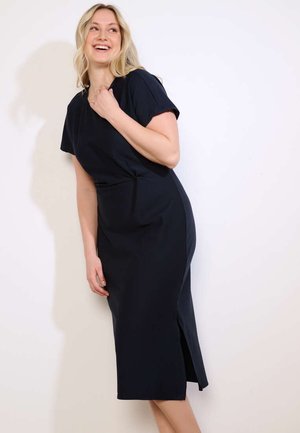 Naine naeratab ja vaatab külili, kandes lühikeste varrukatega põlvpikkust musta kleiti külje lõhikuga, seistes ühevärvse valge tausta ees.