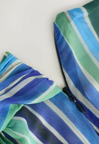 Gros plan d’un tissu rayé bleu, vert et blanc avec une fermeture éclair noire partiellement fermée sur un fond clair.
