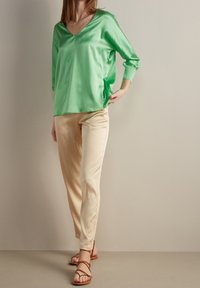 Blouse verte en satin avec un col en V et des manches trois-quarts, associée à un pantalon beige en satin. Le modèle porte des sandales à talons brunes.