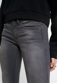 Jeans de denim gris con un corte ajustado, que cuentan con bolsillos delanteros, una cremallera y costuras a tono. Superficie texturizada con un ligero brillo.