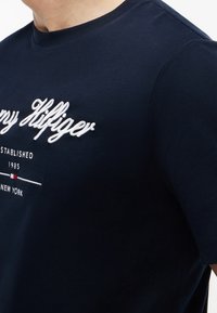 Gros plan d'une personne portant un T-shirt bleu marine avec un logo et un texte Tommy Hilfiger brodés en blanc, incluant "Established 1985 New York."