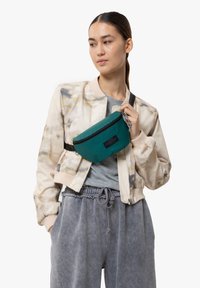 Marsupio teal con cerniera nera e branding, indossato su una leggera giacca bomber sopra una t-shirt grigia e pantaloni della tuta grigi. Texture liscia.