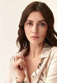 Camisa crema con patrones de hojas marrones, botones en la parte delantera y un cuello. El modelo sostiene el cuello ligeramente abierto, revelando una textura suave.