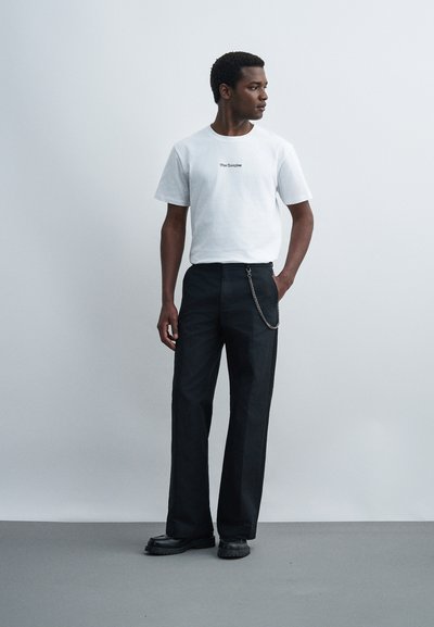 Άντρας όρθιος, φορώντας λευκό t-shirt με μικρό κείμενο, μαύρο παντελόνι με αλυσίδα και μαύρα παπούτσια, κοιτάζει στο πλάι σε απλό φόντο.