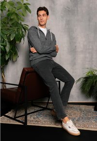 Szary hoodie na zamek błyskawiczny z białymi sznurkami, czarne dżinsy i białe sneakersy na skórzanym fotelu, otoczony zielonymi roślinami i wzorzystym dywanem.