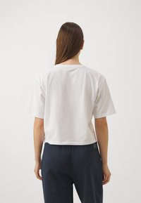Bílý krájený bavlněný tričko s krátkým rukávem a zaobleným výstřihem, vystavené ze zadní strany. Minimalistický design, hladká textura.