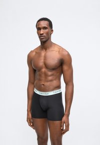 Γυμνασμένος άνδρας όρθιος, φοράει μαύρα εσώρουχα boxer Calvin Klein με λευκή μέση με λογότυπο, σε μονόχρωμο ανοιχτό γκρι φόντο.