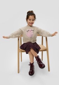 Cremefarbener Pullover mit einem pinken Katzenmotiv, dunkellila Faltenrock und bordeauxroten patentierten Lederstiefeln, sitzt auf einem Holzstuhl.