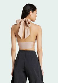 Nude off-shoulder topp med en stor satängrosett vid halsen och strukturerat tyg. Matchas med vida, svarta byxor. Neutral bakgrund.