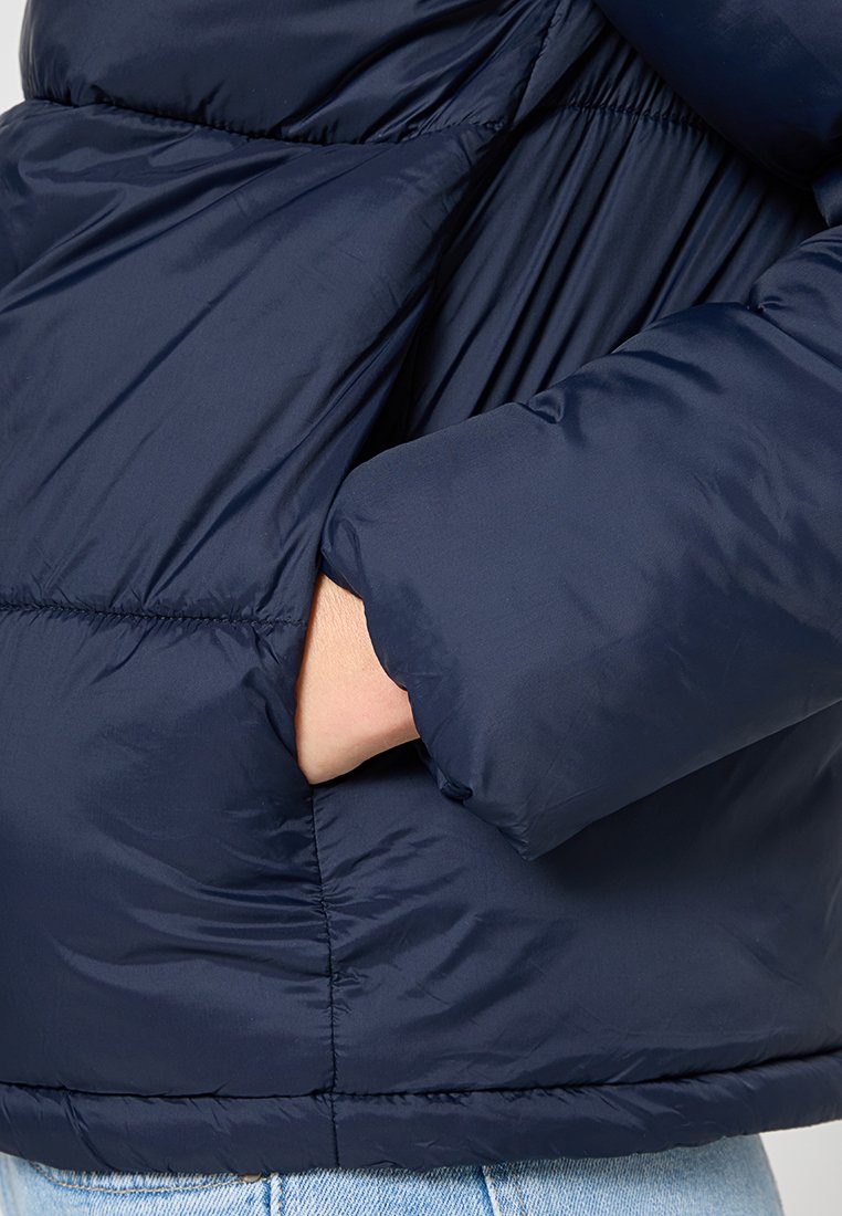 Chaqueta acolchada azul marino con un acabado texturizado, que cuenta con un bolsillo lateral y detalles de tela fruncidos en las costuras y el dobladillo.