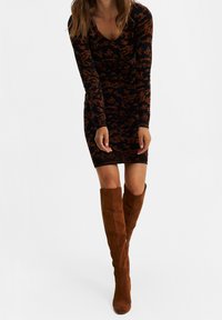 Femme portant une robe longue à manches longues, noire et brune, arrivant au genou, avec des bottes en daim marron montant jusqu'au genou sur fond blanc.