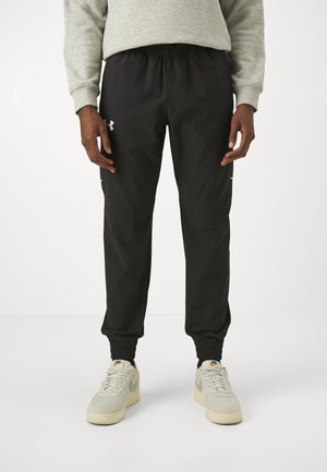 Man die zwarte Under Armour joggers en lichtgrijze Nike sneakers draagt, staand tegen een effen witte achtergrond.