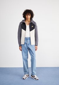 Giacca zip grigia e crema con dettagli neri, abbinata a jeans azzurri chiari e sneakers bianche con dettagli rossi e marroni.