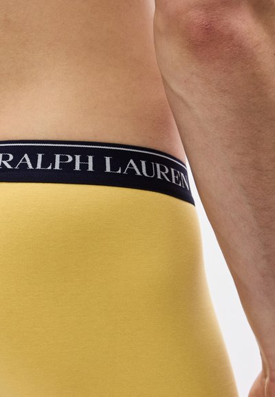 Κοντινό πλάνο μέσης ενός ατόμου που φορά κίτρινο εσώρουχο με μαύρη ζώνη μέσης που φέρει την επιγραφή "RALPH LAUREN" σε λευκό κείμενο.