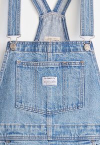 Blå denimsoveraller med justerbare stropper, metalbeslag, forlomme og et syet mærkelabel på brystområdet. Slidt tekstur synlig.